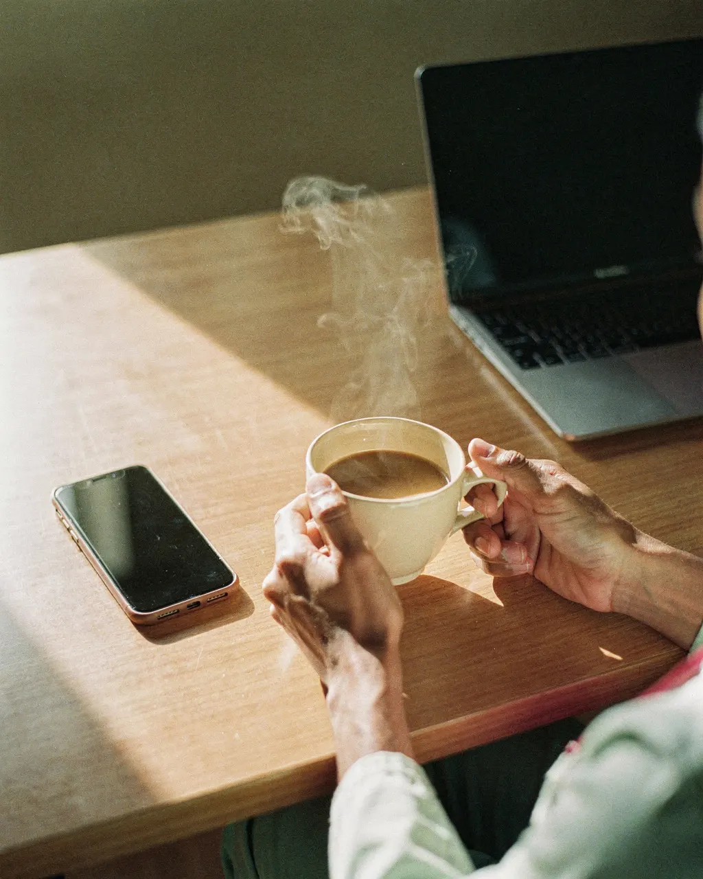 Manos sosteniendo una taza de café caliente junto al celular y la notebook, antes de empezar el día