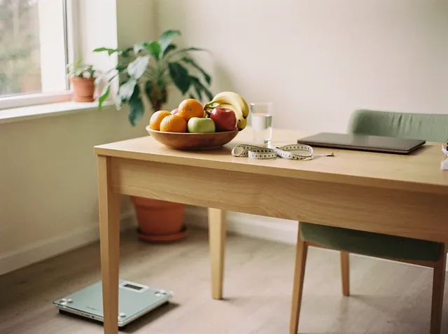 Consultorio de nutrición con un bol de frutas, un vaso de agua, un centímetro y una notebook sobre la mesa de madera, y una balanza en el piso