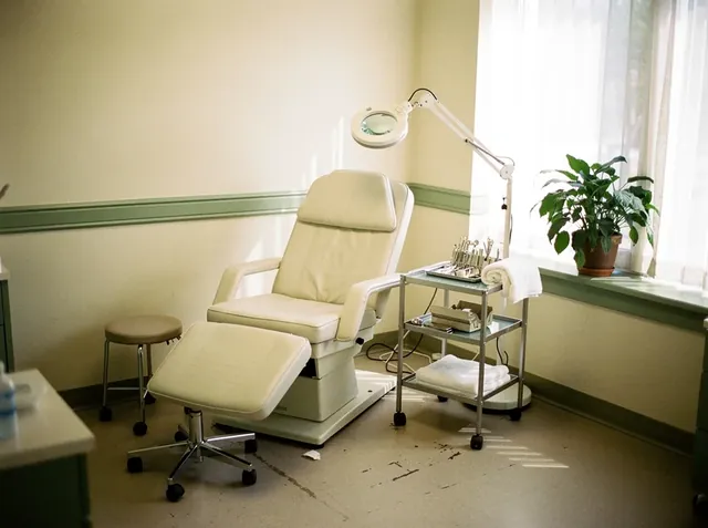Consultorio de podología con sillón reclinable con apoyapiés, lámpara con lupa, banqueta, carrito con instrumental y toallas, y una planta en la ventana