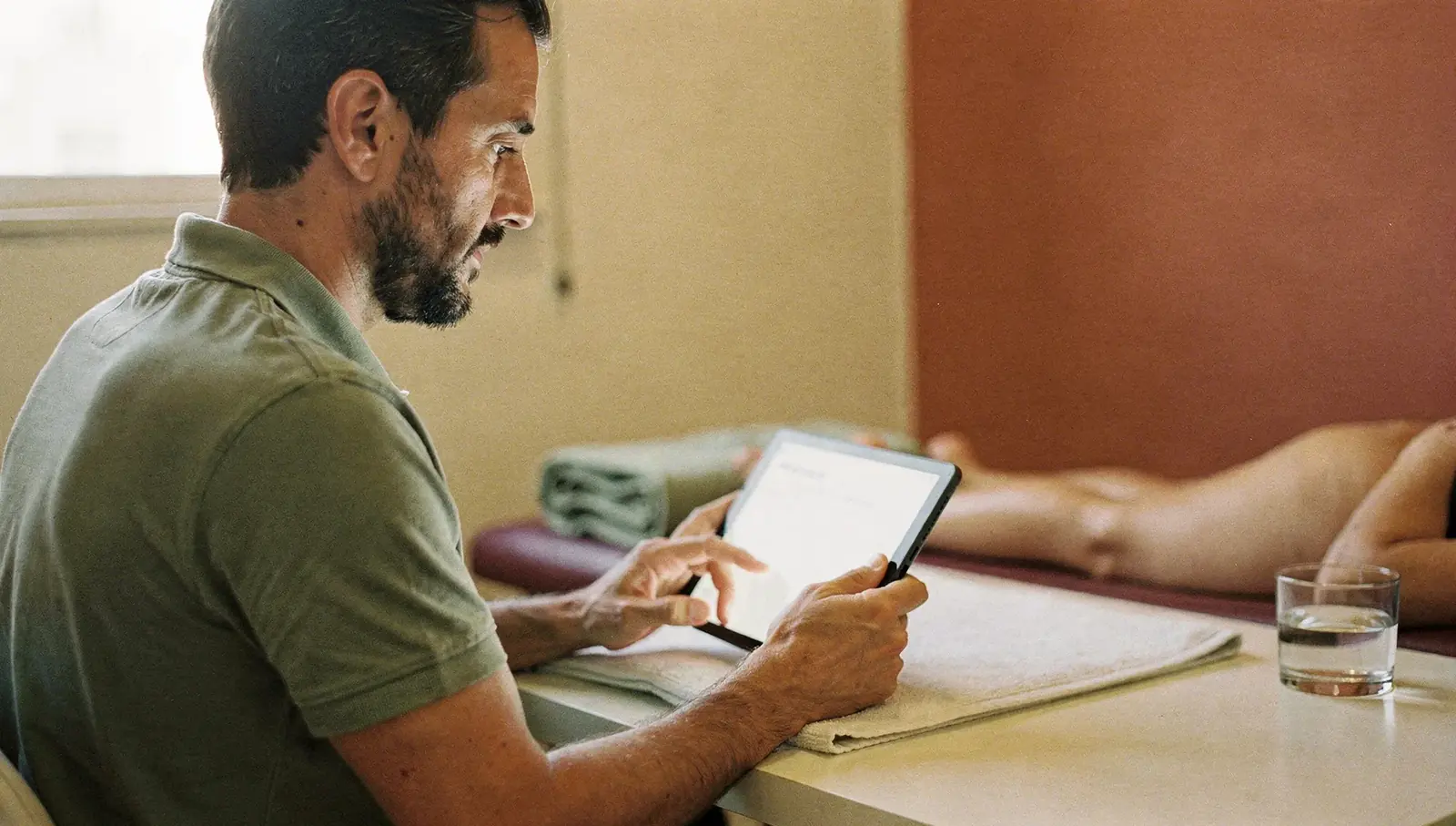 Kinesiólogo con barba completando la evolución de una sesión en una tablet junto a la camilla de su consultorio